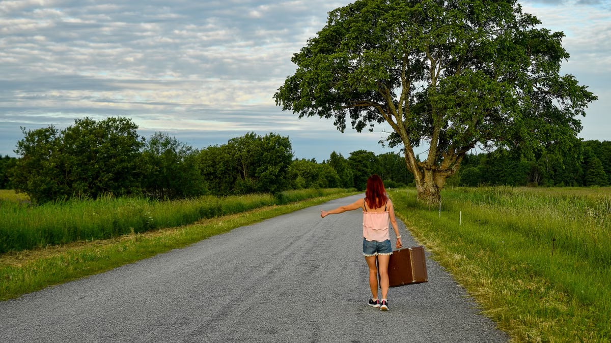 A woman hitchhiking on a picturesque road in Kolga, Estonia, with lush greenery and clear skies.